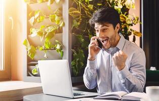 Unbelievable success. Excited millennial businessman in cafe with open laptop, looking at computer screen and talking on phone, empty space photo