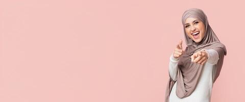 Gotcha. Positive Muslim Girl In Hijab Pointing Fingers At Camera, Posing On Pink Background, Empty Space photo