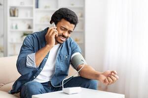 Sick black man sits comfortably in his living room, taking his blood pressure with a modern monitor, as he has a conversation with his doctor on his smartphone photo