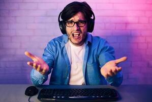 A young man sits at a computer desk, wearing headphones and looking frustrated with his hands raised in the air. He is likely reacting to a difficult game play or an unexpected loss. photo