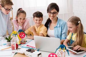 Elementary school students are working on a robotics project in their classroom. The students are gathered around a laptop and are looking at the screen intently. Their teacher is sitting beside them photo