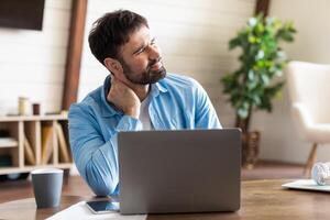 A man is sitting at a wooden table, focused on his laptop with a pained expression, rubbing the back of his neck. The setting appears to be a bright and cozy home office photo