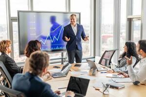 A group of professionals are gathered around a table in a modern office, listening to a businessman who is standing in front of a large screen displaying a graph showing upward growth. photo