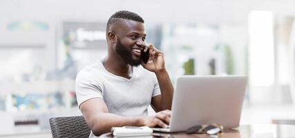 A man in a white t-shirt smiles while talking on the phone and using a laptop in an office setting. photo