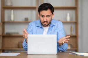 A man sits at a desk with an expression of confusion or surprise as he stares at his laptop screen, with papers scattered around him suggesting he is in the midst of work photo