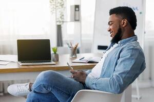 Idleness in office. Carefree black employee using smartphone during working hours browsing internet or typing message, side view photo
