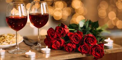 Romantic Dinner. Bouquet of flowers lying on the table, selective focus on bunch of roses, two glasses of red wine and candles on the wooden desk. Date concept, blurred background, banner, copy space photo
