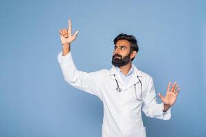 A bearded Indian doctor wearing a white lab coat and stethoscope gestures with his right hand as if pointing towards something, set against a solid blue background. photo