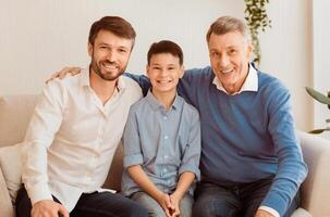 Smiling Boy With Father And Grandfather Sitting On Sofa Hugging And Looking At Camera At Home. Male Generation Family Portrait. photo