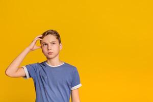 Pensive teenage boy looking up with thoughtful expression, holding hand on head, trying to remember something, orange background with free space photo