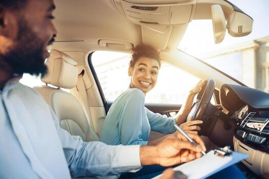A driving instructor sits in the passenger seat of a car while a student driver sits in the drivers seat and turns to look at the instructor. The instructor is using a pen to write on a clipboard. photo