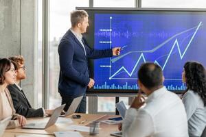 A businessman in a suit points to an upward trending line graph on a large screen while presenting to his team during a meeting. His team sits around a table with laptops open photo