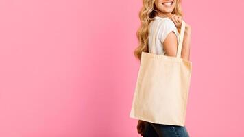 A woman with long blonde hair smiles and holds a white canvas tote bag against a pink background. Her body is turned to the side, and she is only partially visible in the photo, cropped photo
