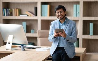 A smiling Indian businessman wearing a blue blazer stands in a modern office, leaning against a desk with a computer monitor in the background. He is holding a tablet in his hands photo
