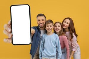 A cheerful family of four is posing closely together with the father extending his arm to show a smartphone with a blank white screen, suggesting an interactive presentation or a mockup. photo