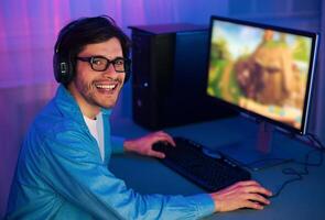 A young man with a blue shirt and headphones plays a game on a computer at night. He is smiling and appears to be having a good time. The computer screen is lit up with the colors of the game photo