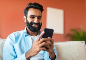 Portrait Of Cheerful Indian Man Using Mobile Phone Surfing Internet, Networking And Texting Sitting On Sofa Indoors. Young Bearded Guy Messaging And Communicating Via Smartphone Gadget Online photo