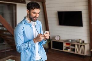 A man is standing in a living room, focused on his cell phone screen. He appears engaged in whatever he is observing or reading on the device. photo