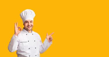 Smiling Chef Man Gesturing Okay Pointing Finger At Copy Space Approving Something Posing Over Yellow Studio Background. Panorama photo
