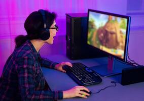 This image shows a young woman wearing a plaid shirt and headphones, playing a game on a computer. She is sitting at a desk with a keyboard and mouse. photo