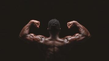 A muscular man flexes his biceps, showcasing his strength and physique. The image is taken from behind against a black backdrop. photo
