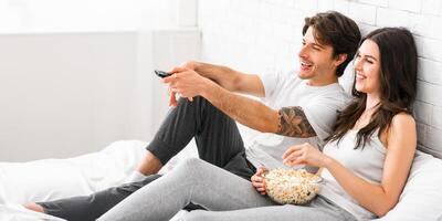 A man and woman are relaxing on a bed, watching television. They are both smiling and laughing, enjoying each others company. The man is holding a remote control and the woman is eating popcorn photo