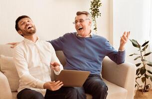 Cheerful Middle-Aged Son And Senior Father Using Laptop And Laughing Sitting On Couch At Home. Selective Focus photo