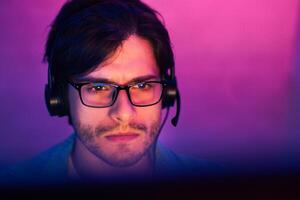 This image shows a young man with dark hair wearing glasses and headphones while looking at a computer screen. He has a serious expression on his face, suggesting he is focused on his task. photo
