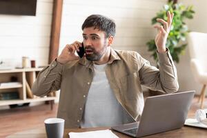 Man sits at wooden table, showing signs of frustration during phone conversation. His laptop is open, positioned in front of him, and he gestures with one hand, expressing his annoyance or confusion photo