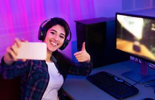 This image shows a woman sitting at a desk with a computer and keyboard. She is wearing headphones and taking a selfie with her smartphone. She is smiling and giving a thumbs-up. photo