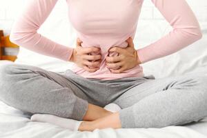 A woman is seen sitting on a bed in a room, holding her stomach with both hands. She appears to be in discomfort or pain, possibly experiencing abdominal cramps or digestive issues. cropped photo
