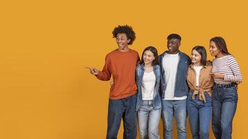 A group of five friends stand together in front of a bright yellow background. They are smiling and looking happy, with one person pointing to the side at copy space photo