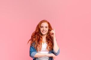 Cheerful young woman with flowing red hair is standing against a pink backdrop, pointing upward with one finger, her expression is friendly, appears to be engaging with someone or presenting an idea. photo