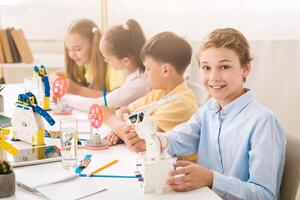A young boy smiles at the camera while holding a robot during a STEM class. He is sitting at a table with other children who are also working on STEM projects. The classroom is bright and airy photo