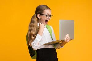 Computer Game Addiction. Overjoyed School Girl Playing Computer Games On Laptop Over Yellow Background In Studio. Free Space For Text photo