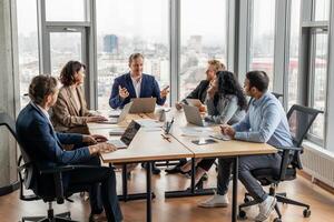 A group of business professionals are gathered around a large conference table in a modern office, engaging in a discussion. Laptops and documents are spread out on the table photo