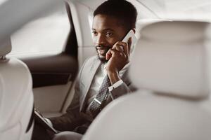 African American man manager in a suit sits in the backseat of a car and talks on his phone. He looks intently at the phone and appears to be engaged in a conversation. photo
