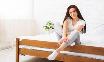 A woman is seated on top of a wooden bed, looking towards the camera. The room is minimally decorated, with the bed being the central focus. photo