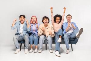 A group of diverse young adults are excitedly celebrating together on a couch. They are all smiling and laughing, with their arms raised in the air. photo