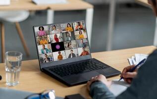 A person sits at a desk in an office and participates in a conference meeting on their laptop computer. The laptop screen displays a grid of multiple participants. photo