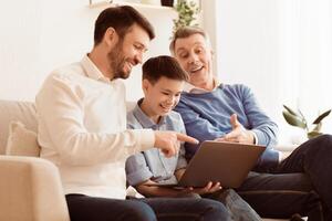 Happy Boy Using Laptop Sitting Between Father And Grandfather, Laughing And Having Fun Together On Couch At Home. Selective Focus photo