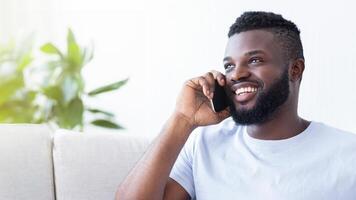 A happy, smiling black man with a beard is talking on the phone while sitting on a white couch. He is wearing a light blue t-shirt and is looking off to the side, copy space photo