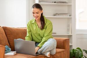 A woman is sitting comfortably on a couch, focused on using a laptop computer. She appears engaged in her digital tasks, with a concentrated expression. The room is well-lit photo