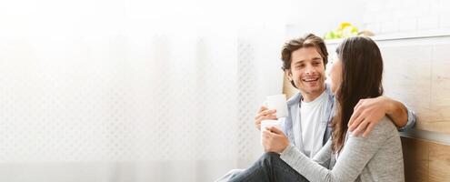 A happy couple sits on the floor of their kitchen, engaging in a conversation and enjoying each others company. The man is holding mug, while the woman leans her head towards him with a gentle smile. photo