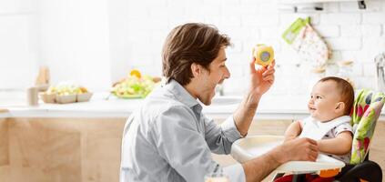 A father sits in a kitchen with a baby in a high chair. He is holding a yellow toy and making the baby laugh. The kitchen is bright and clean. photo