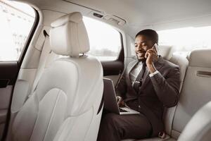 Black businessman in a suit sits in the backseat of a car. He is talking on his phone and using a laptop computer, appears to be in professional setting and is likely on his way to important meeting. photo
