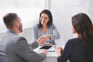 A woman in a gray blazer smiles and shakes hands with a man sitting across from her at a table. A third person sits to the woman right and observes the interaction. photo