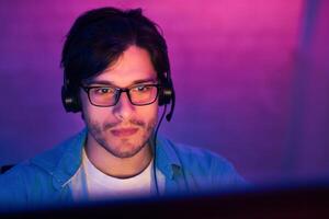 A young man with short, dark hair, wearing glasses and a headset, looks intently at a computer screen. He has a serious expression on his face. The background is blurred photo