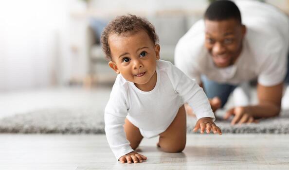 linda pequeño negro infantil bebé gateando en piso a hogar, orgulloso joven padre mirando a él y sonriente, papá y niñito niño disfrutando gasto hora juntos, selectivo atención con Copiar espacio foto