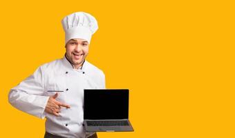 Chef Guy Showing Laptop With Empty Screen Standing Over Yellow Studio Background. Culinary Blog. Mockup, Panorama photo